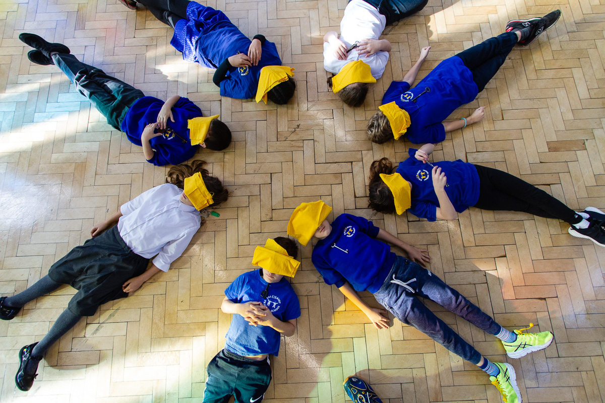 Children laying on floor in a circle with heads together, faces covered with yellow clothes, covered in dappled sunlight.