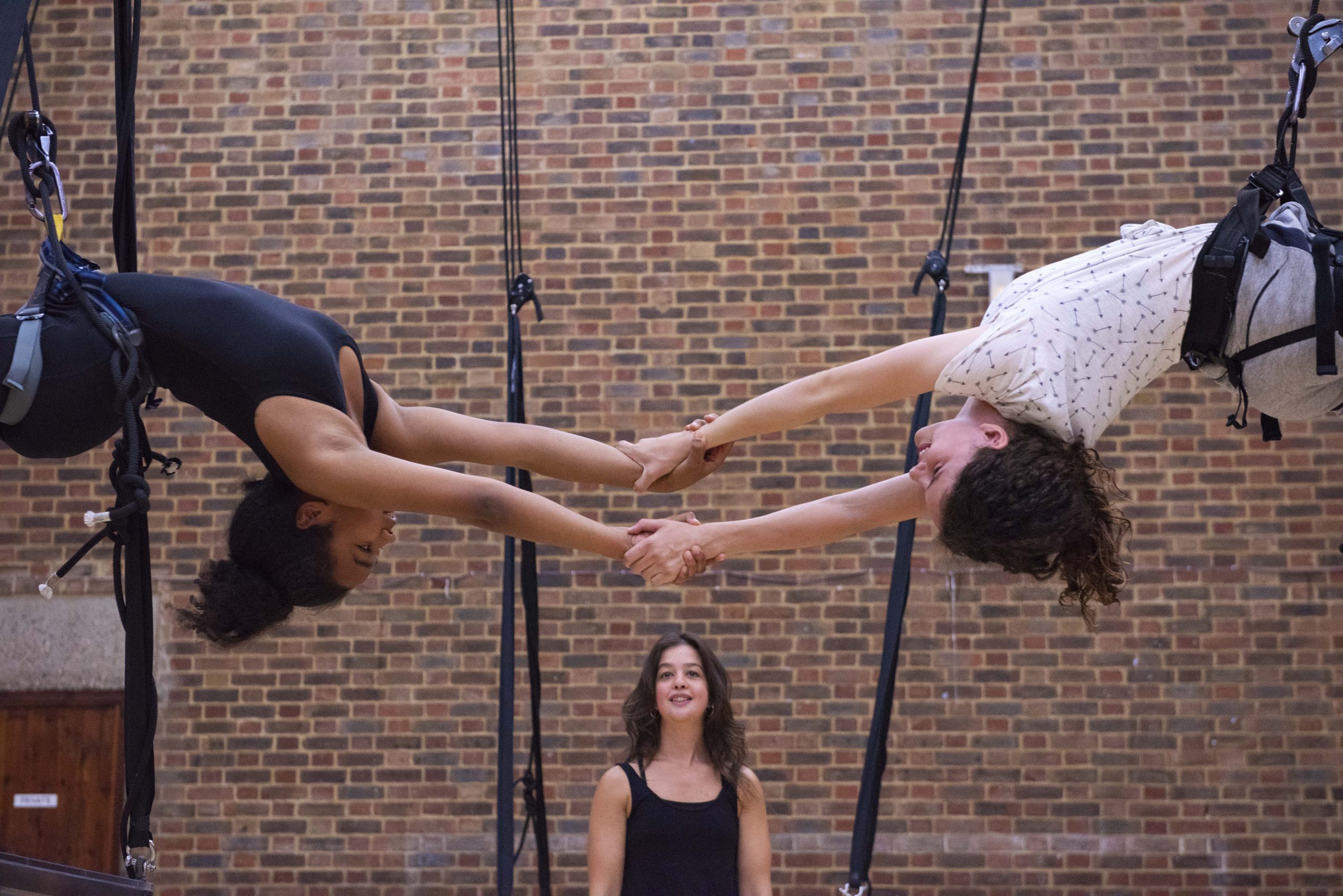 Two young aerialists in harnesses are suspended in the air, they clasp each others hands to link them across the image. Below another person stand in the middle underneath them smiling up at them.