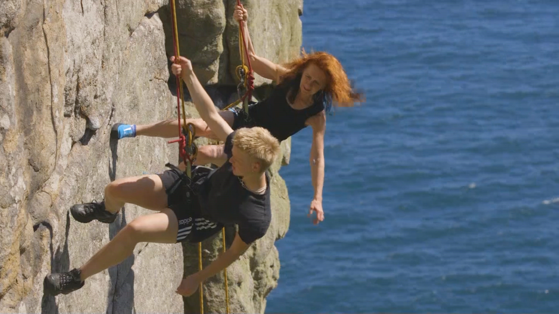 On a sunny day mother and son abseil down the side of golden Cornwall cliffs with a bright blue sea in the background