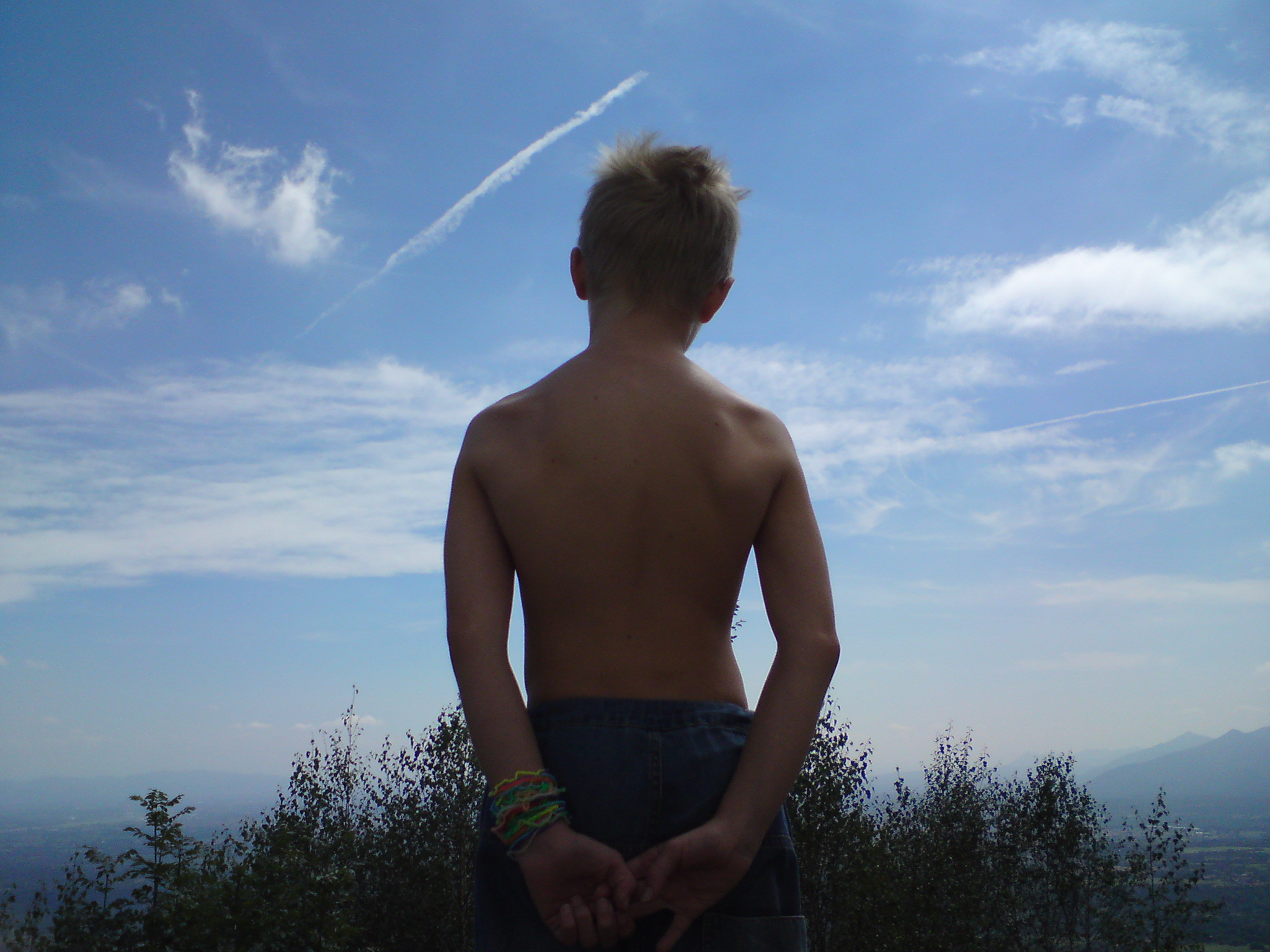 Emerging. A child stands with their bare back to the camera, looking out at a blue sky. They have short blond hair and are holding their hands behind them. Their lower half is obscured by long grass which is in shadow