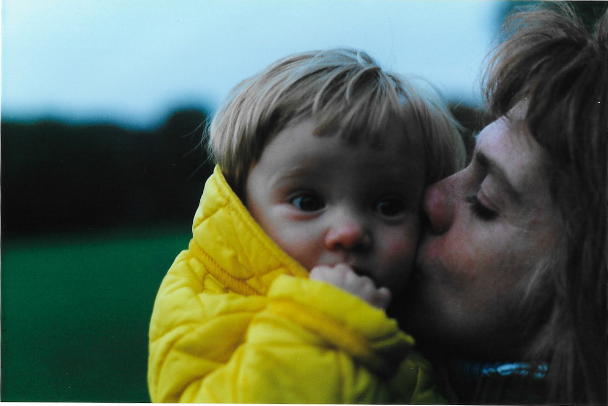 A mother kisses the cheek of her child. The child is wearing a bright yellow coat and has ruddy cheeks and nose.