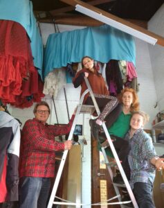 Emma sits on the top of a ladder, two people stand on the ladder steps as another supports the back of it.