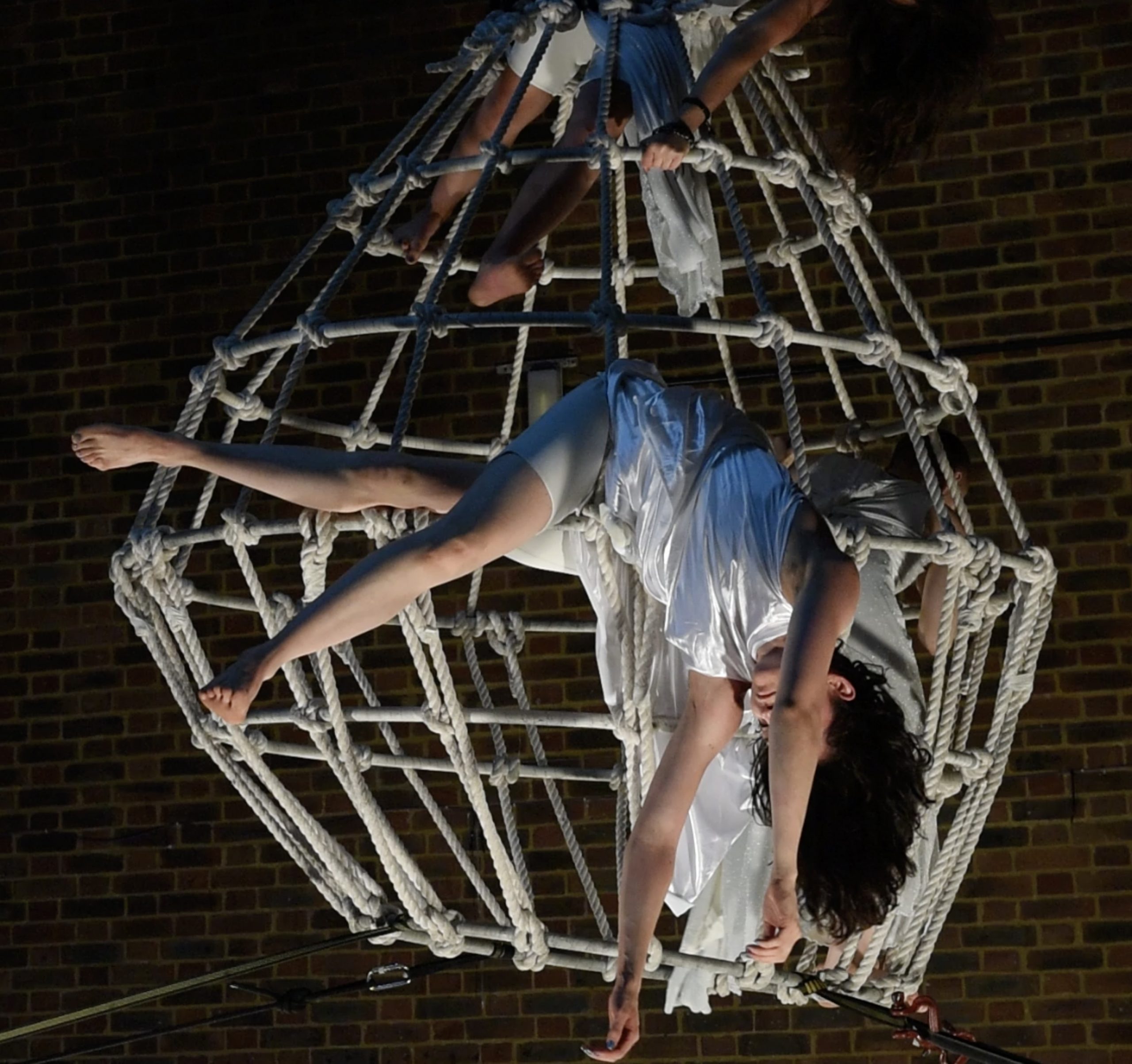 Performers in 'Fall to Fly', woman in white flowing costume suspended from a harness over white rope cage structure.