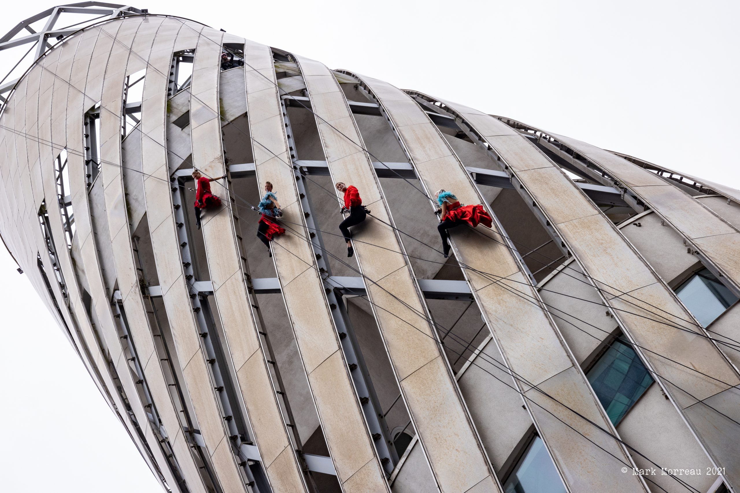 Four aerial performers in red costumes suspended on the side of The Lowry building.