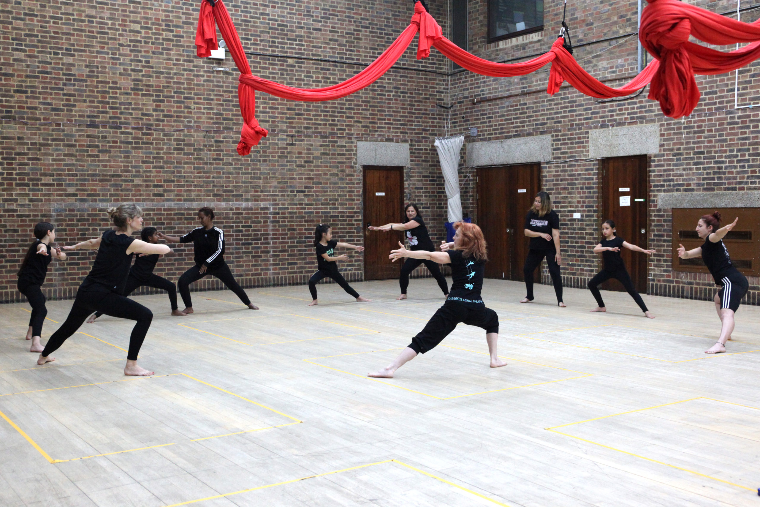 A group of families and tutors warm up in a circle, stretching their arms, the bright red silks suspended above their heads contrast with the pale wooden floor of the hall they're in.