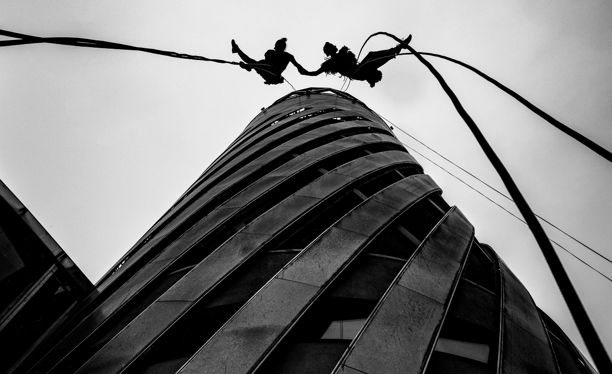 Black and white photograph of two performers suspended in front of large spiral building