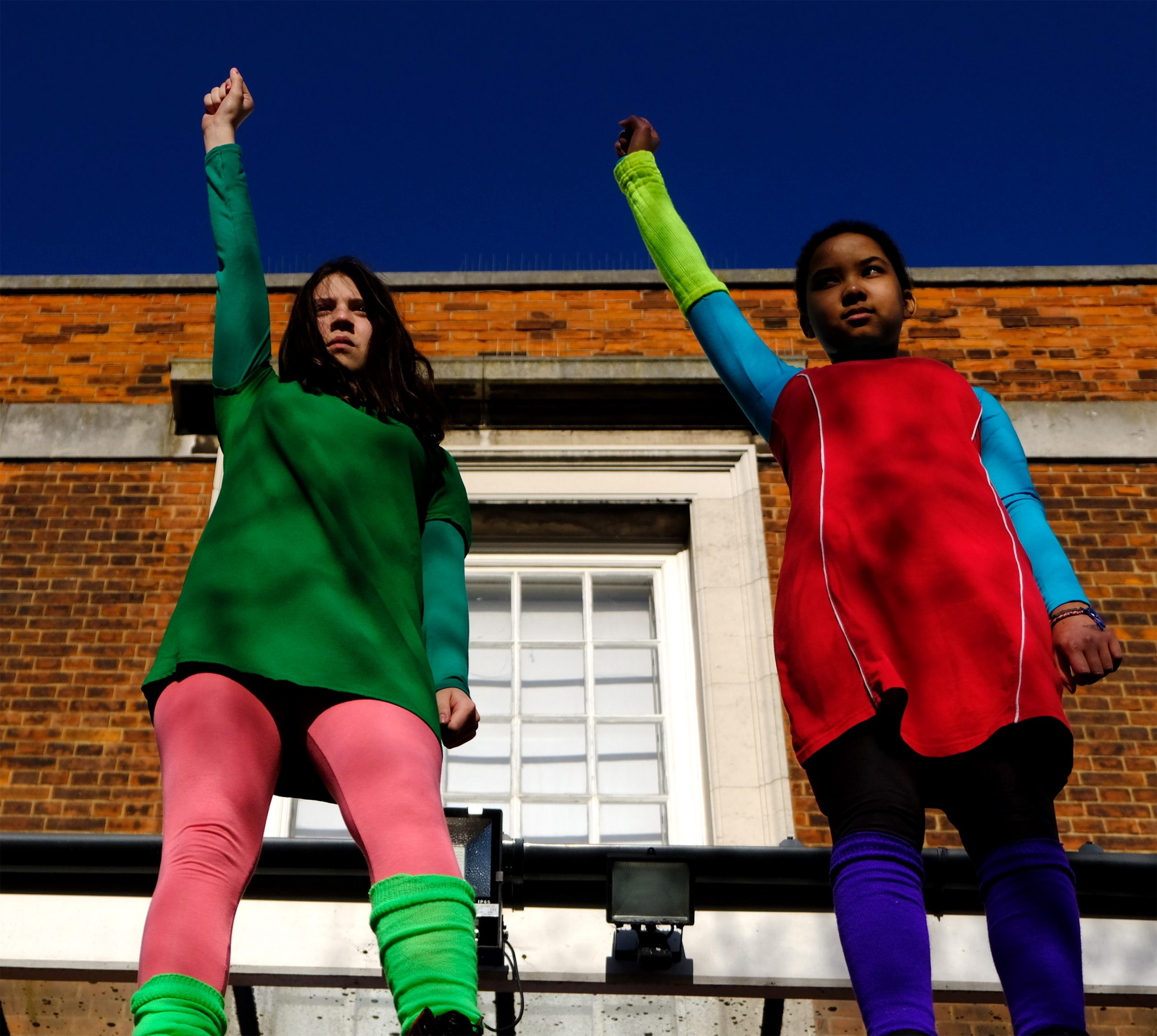 Two young students in brightly coloured clothes raise a closed fist and arm into the air in solidarity.