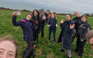 A group of people take a selfie while standing in a field on the top of a cliff, we can see an overcast sky and skyscape in the background