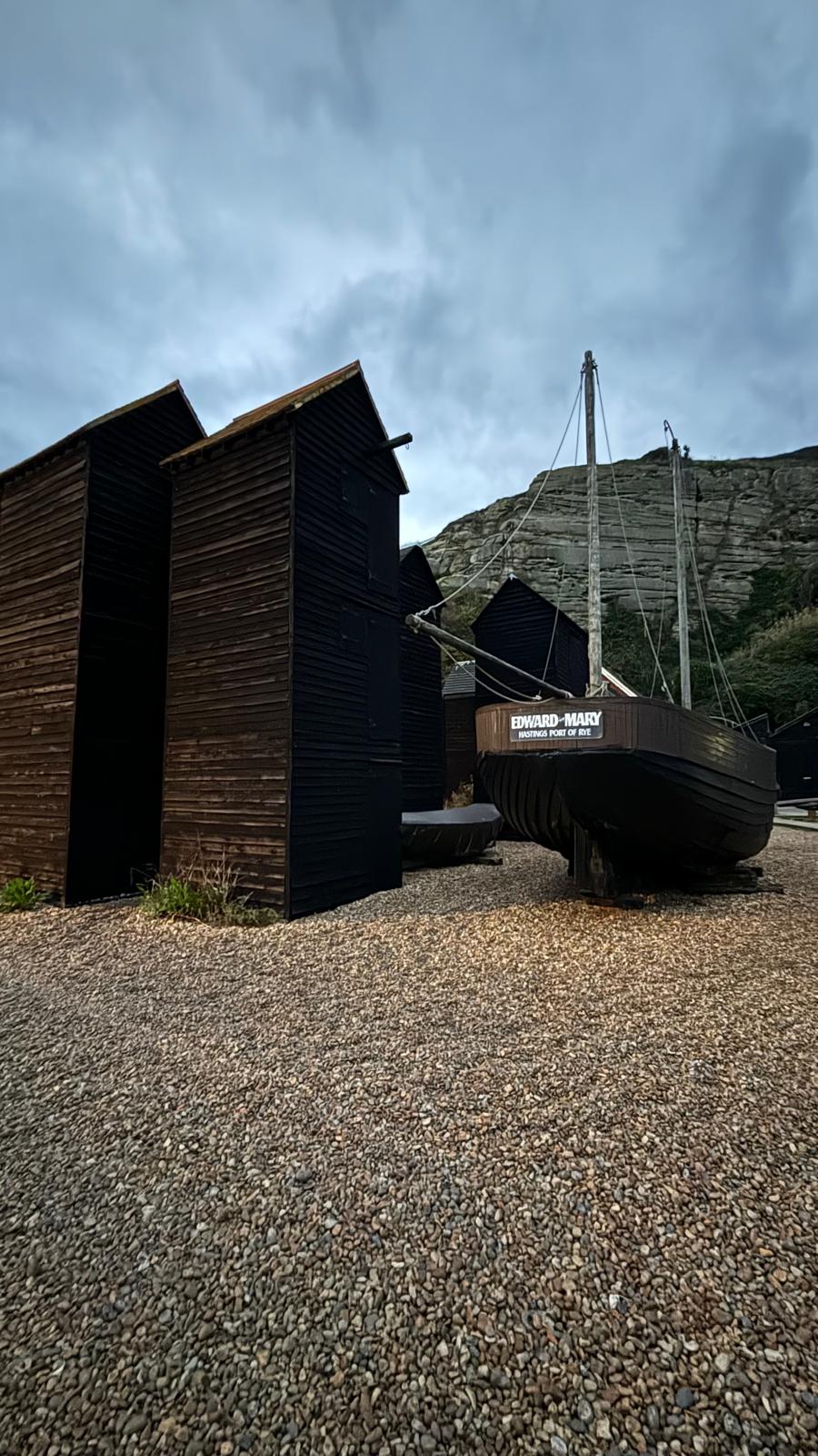 Two tall black fishing huts stand next to a beached boat. They sit on pebbles with a cloudy blue sky behind them